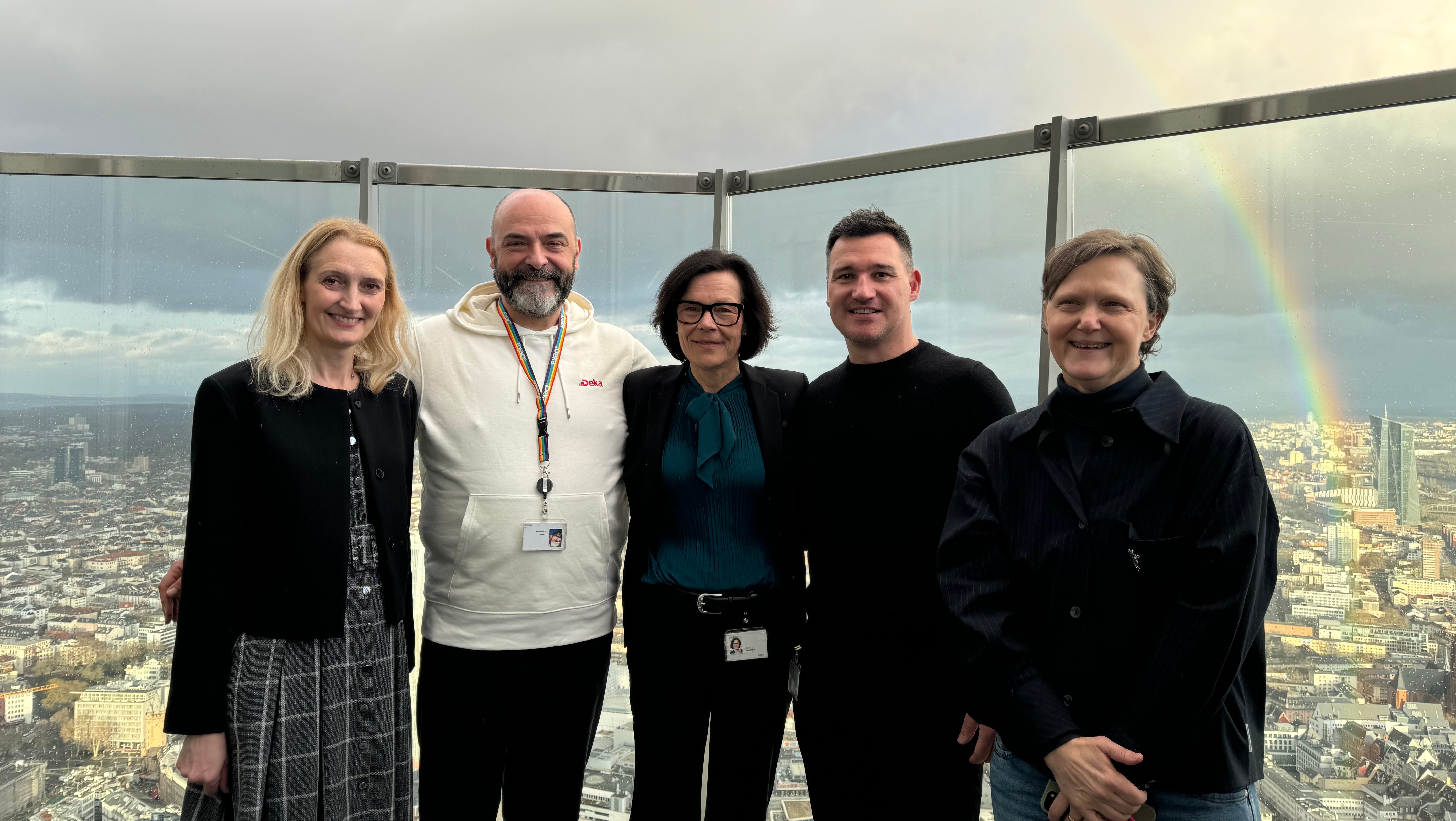 Drei Frauen und zwei Männer stehen auf Dachterrasse mit Regenbogen im Hintergrund